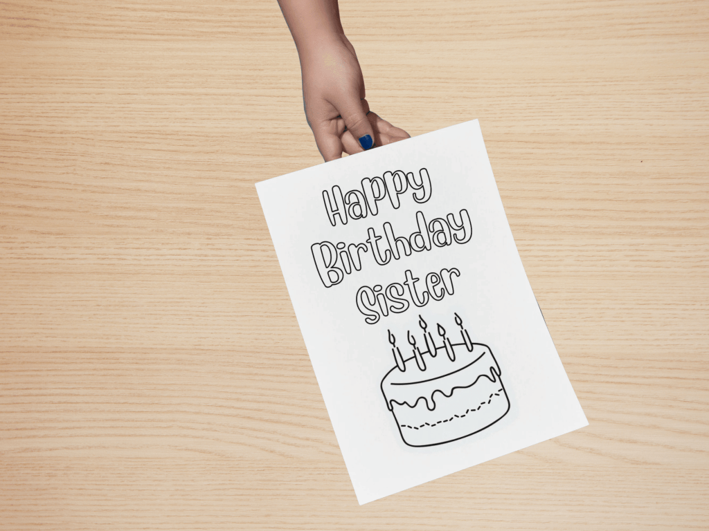A child’s hand holding a birthday card on a wooden table. The card reads "Happy Birthday Sister" above a drawing of a cake with candles.