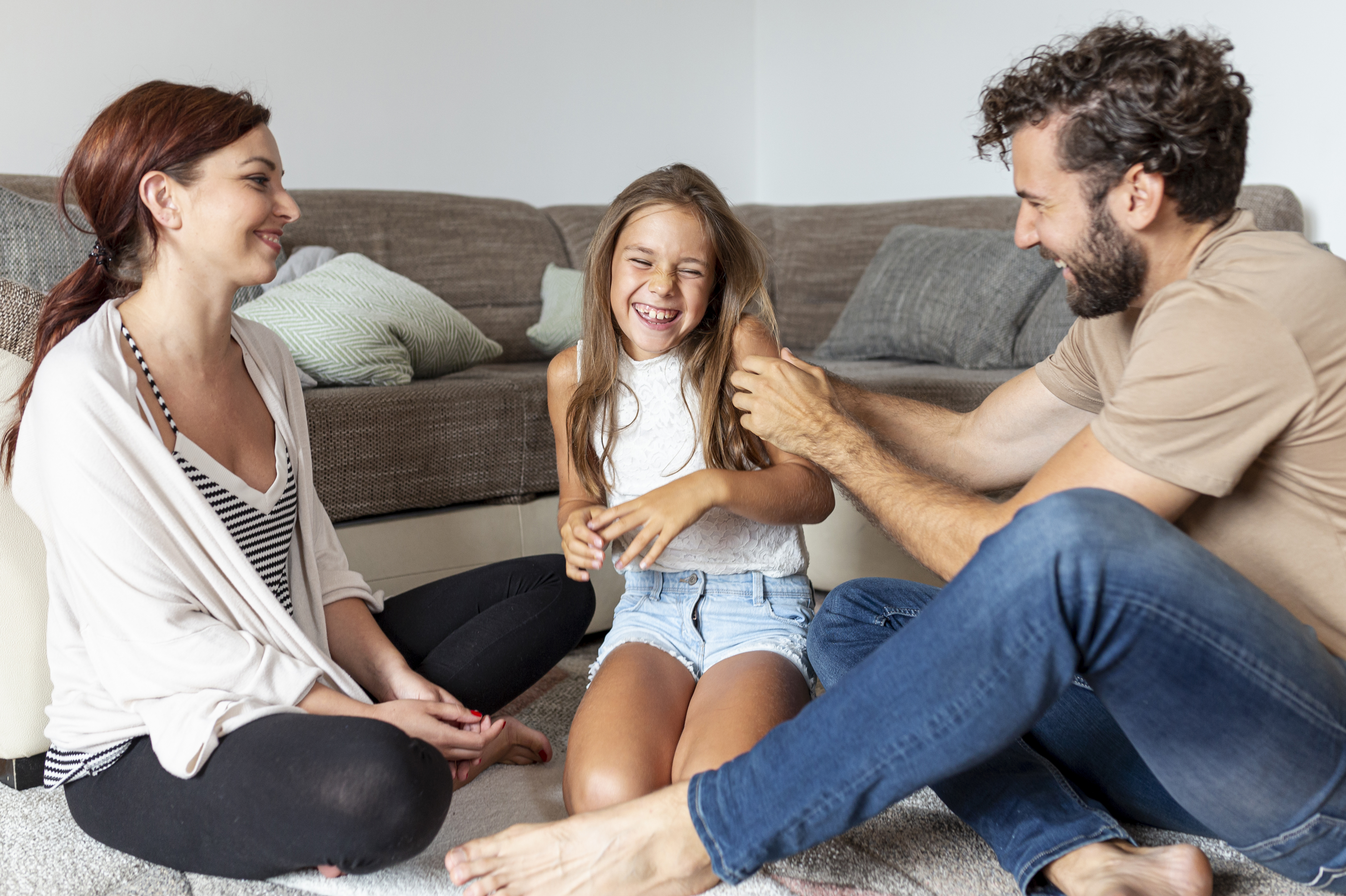 A family enjoys quality time on the floor; a girl laughs energetically as her parents, sitting cross-legged, playfully interact with her in a cozy living room.